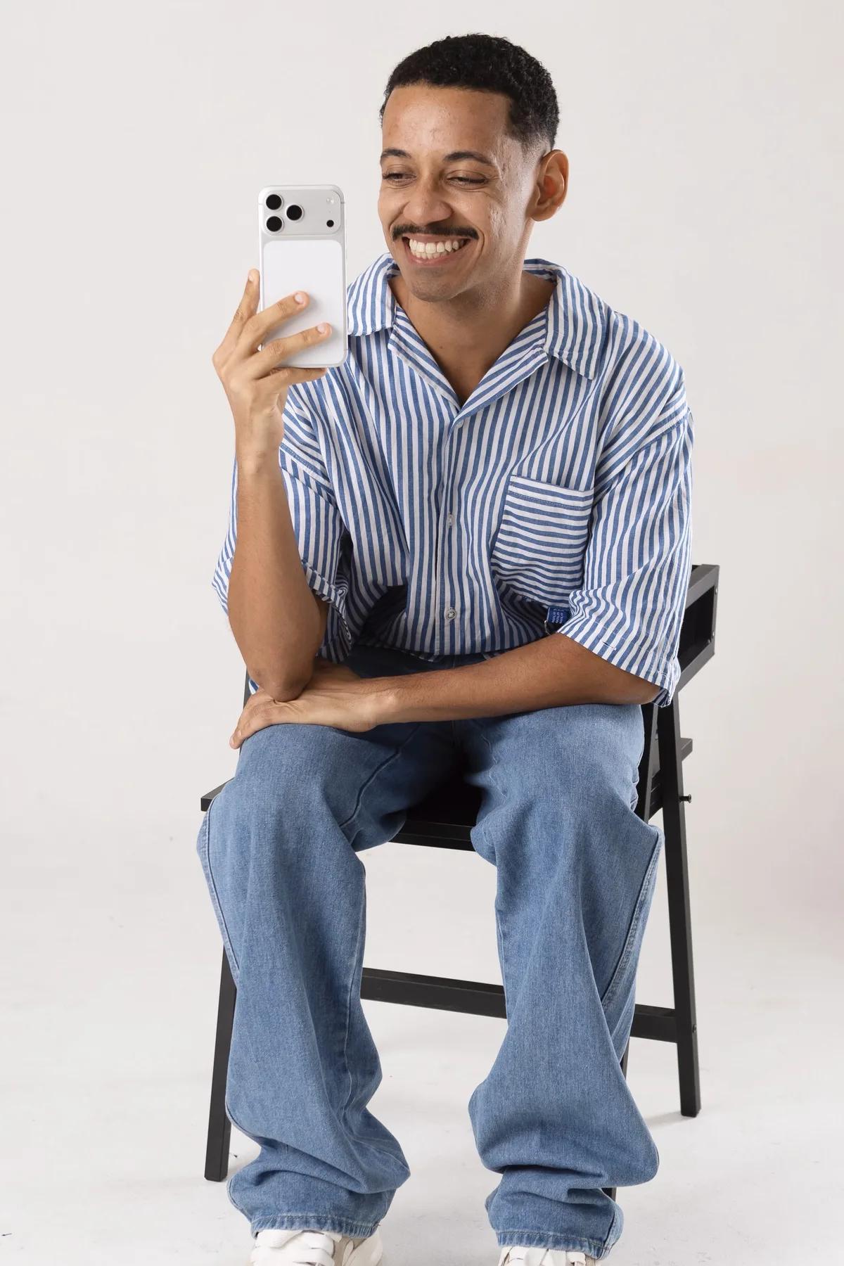 Yugui Alves sorrindo, sentado em estúdio de fundo branco, segurando o celular